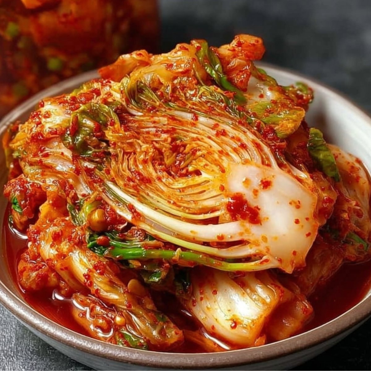 Bowl of freshly made Fresh Kimchi featuring napa cabbage in a spicy red pepper paste, with a jar of kimchi in the background on a dark surface.