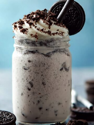 Glass jar of Oreo milkshake topped with whipped cream, crushed Oreos, and a whole Oreo cookie, with a black straw and cookies scattered around.