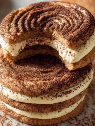 Two stacked soft cookies dusted with cocoa powder, filled with creamy mascarpone-style filling. The top Tiramisu Cookies has a visible bite taken out, showing the soft interior. Inspired by tiramisu, these sandwich cookies blend dessert and espresso flavors.