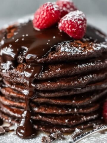 Stack of rich chocolate pancakes topped with melted chocolate, powdered sugar, and fresh raspberries on a white plate