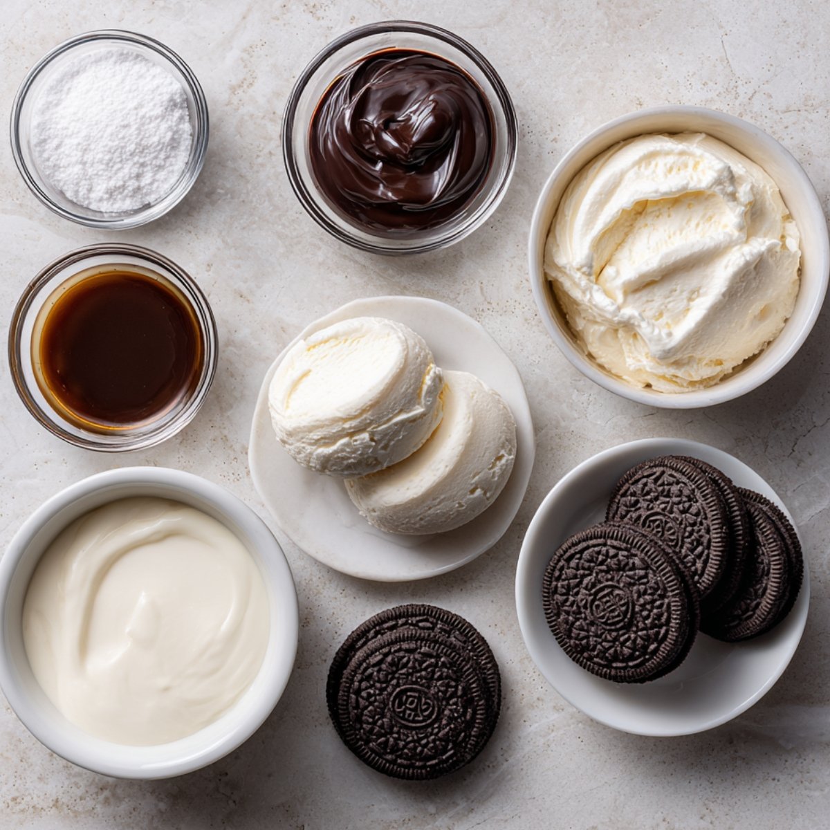 Ingredients for an Oreo milkshake on a white marble surface, including whipped cream, Oreos, vanilla extract, powdered sugar, chocolate sauce, ice cream scoops, and yogurt