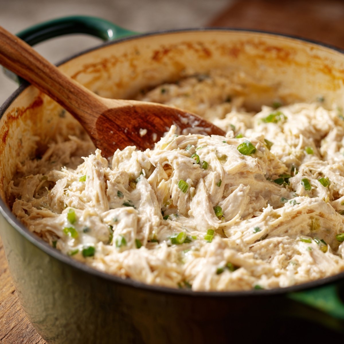 Shredded rotisserie chicken mixed with cream cheese and chopped green onions in a green Dutch oven.