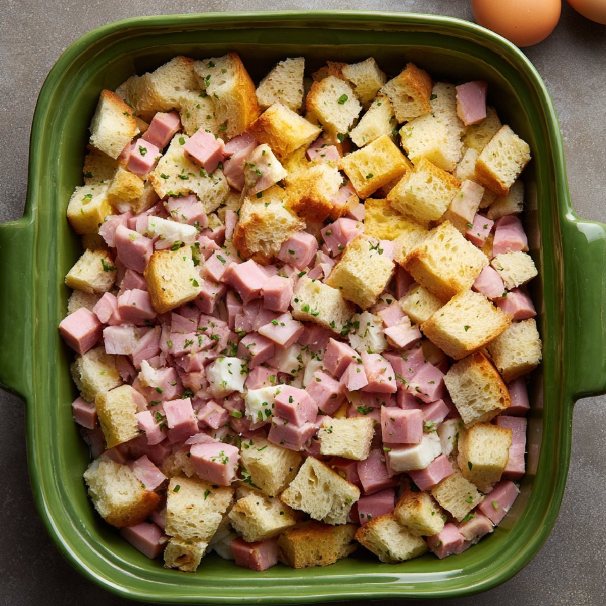 Overhead view of a green baking dish filled with cubed bread and diced ham, prepared as the base layer for an eggs Benedict casserole.