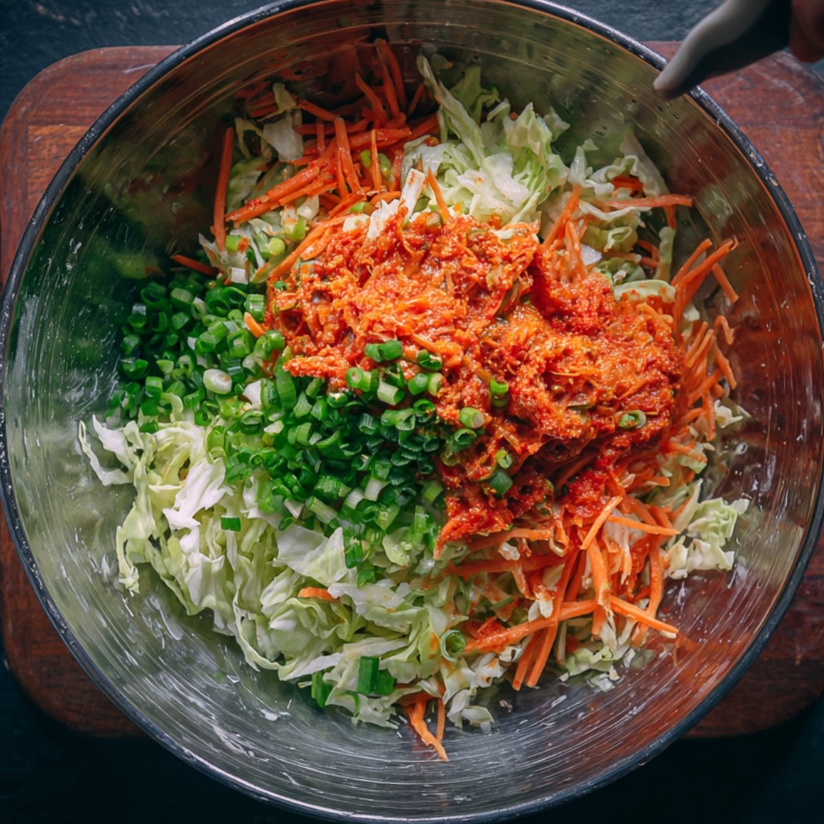 Large metal mixing bowl filled with shredded napa cabbage, carrots, chopped green onions, and a generous scoop of red kimchi paste, ready to be mixed by hand.