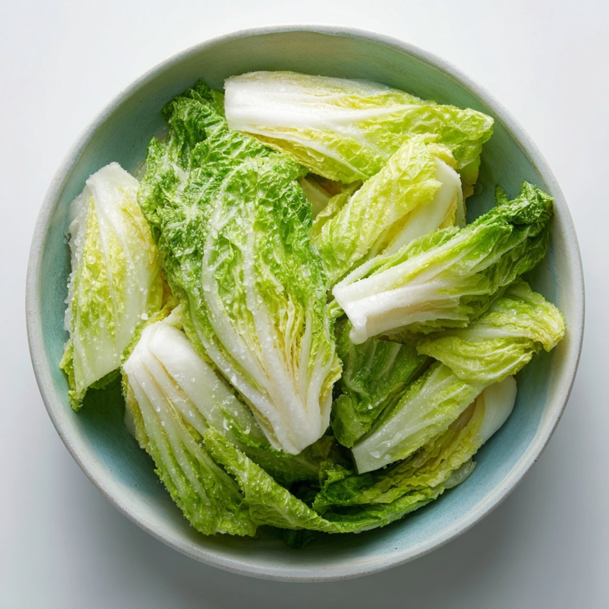 Light green napa cabbage leaves in a ceramic bowl, visibly coated in coarse salt, soaking and wilting in preparation for kimchi.