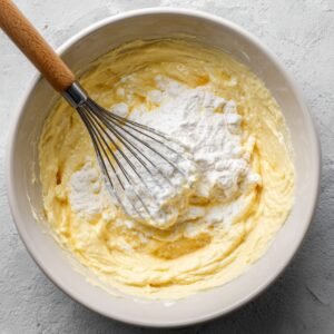 Mélange à pancakes dans un bol, avec de la farine ajoutée sur le dessus et un fouet en bois prêt à incorporer les ingrédients.