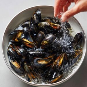 Fresh mussels being rinsed in a bowl of cold water, with a hand sprinkling water over them.