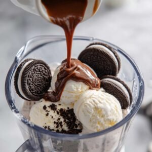 Close-up of a blender with scoops of vanilla ice cream, whole Oreo cookies, and chocolate syrup being poured on top, ready to be blended into a milkshake.