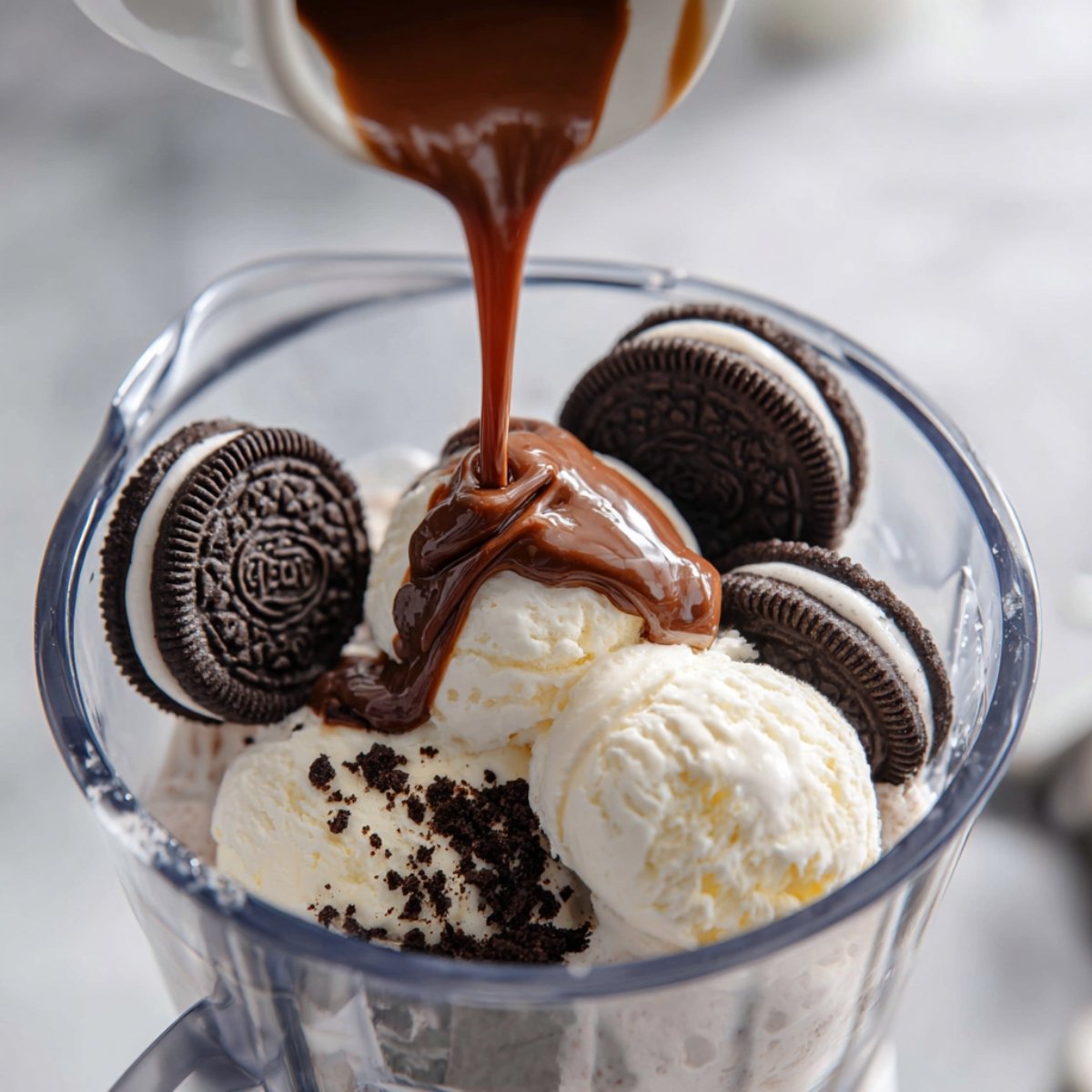 Close-up of a blender with scoops of vanilla ice cream, whole Oreo cookies, and chocolate syrup being poured on top, ready to be blended into a milkshake.