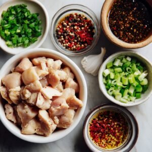 Overhead view of raw diced chicken surrounded by small bowls of chopped green onions, cracked pepper, soy sauce, chili oil, and garlic cloves, arranged on a white marble surface.