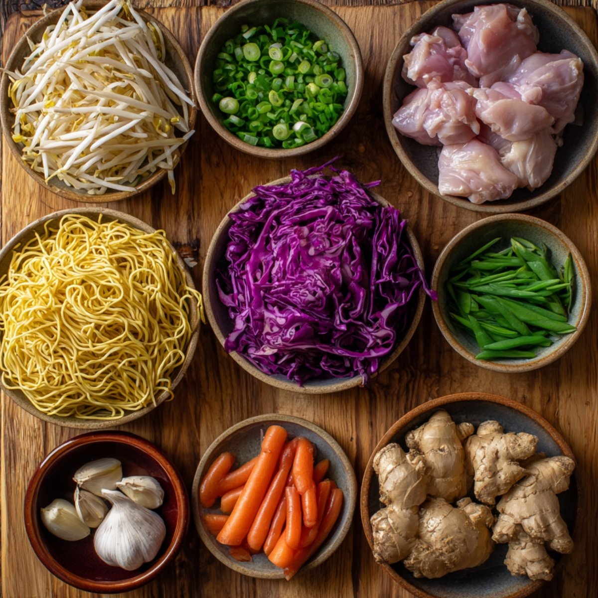 A wooden board displaying fresh ingredients for chicken chow mein: raw chicken pieces, chow mein noodles, shredded purple cabbage, bean sprouts, chopped scallions, sliced green beans, carrots, garlic, and ginger.