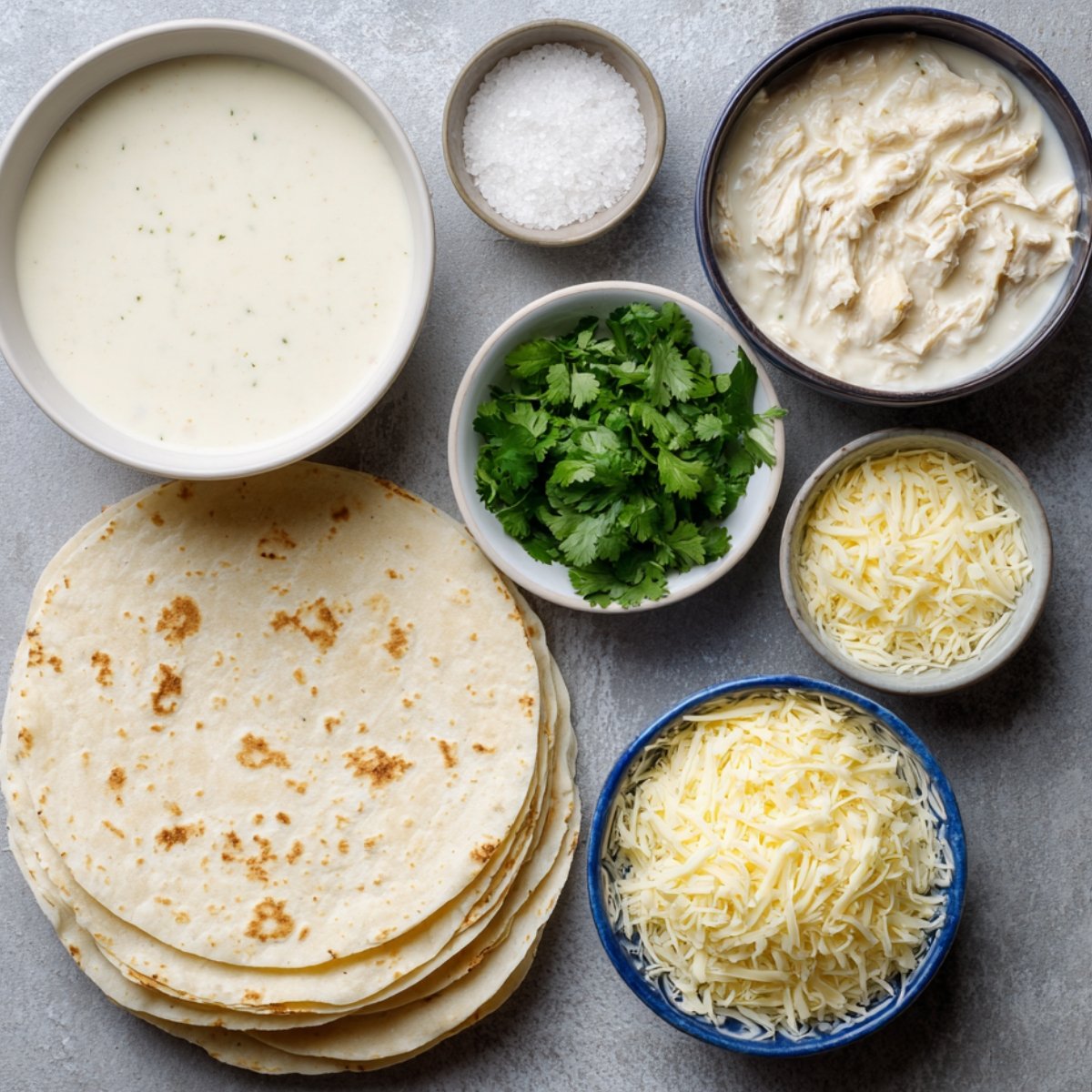 Tortillas, shredded cheese, creamy chicken filling, white sauce, and fresh cilantro arranged in bowls.