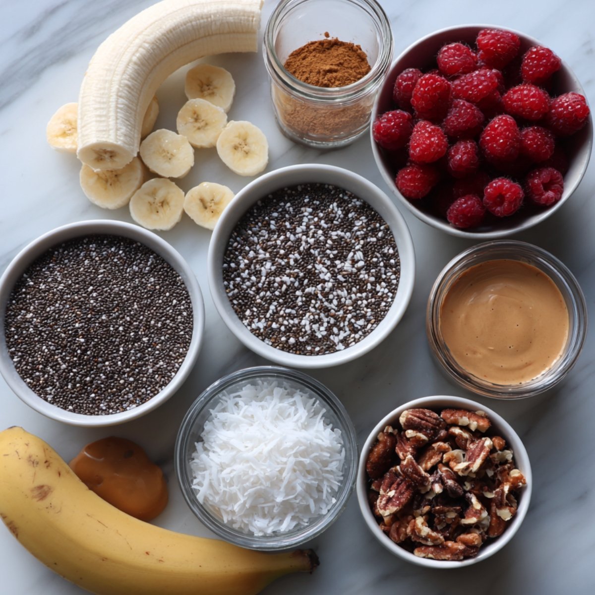 Top view of chia seed pudding ingredients including bananas, raspberries, chia seeds, shredded coconut, peanut butter, pecans, cinnamon, and coconut flakes arranged on a marble surface.